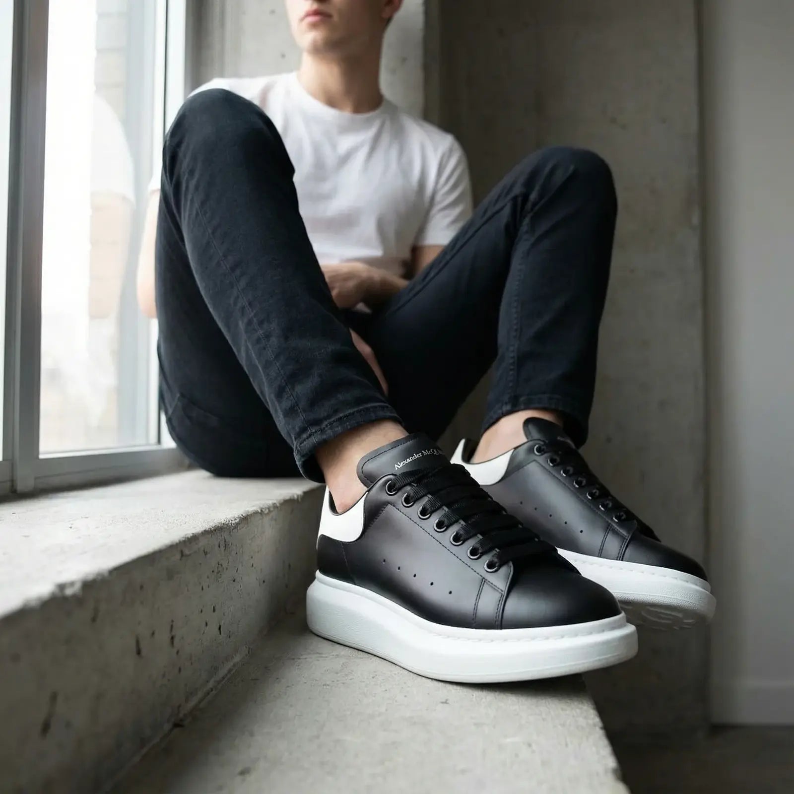 Person wearing black sneakers with white soles sitting by a window.