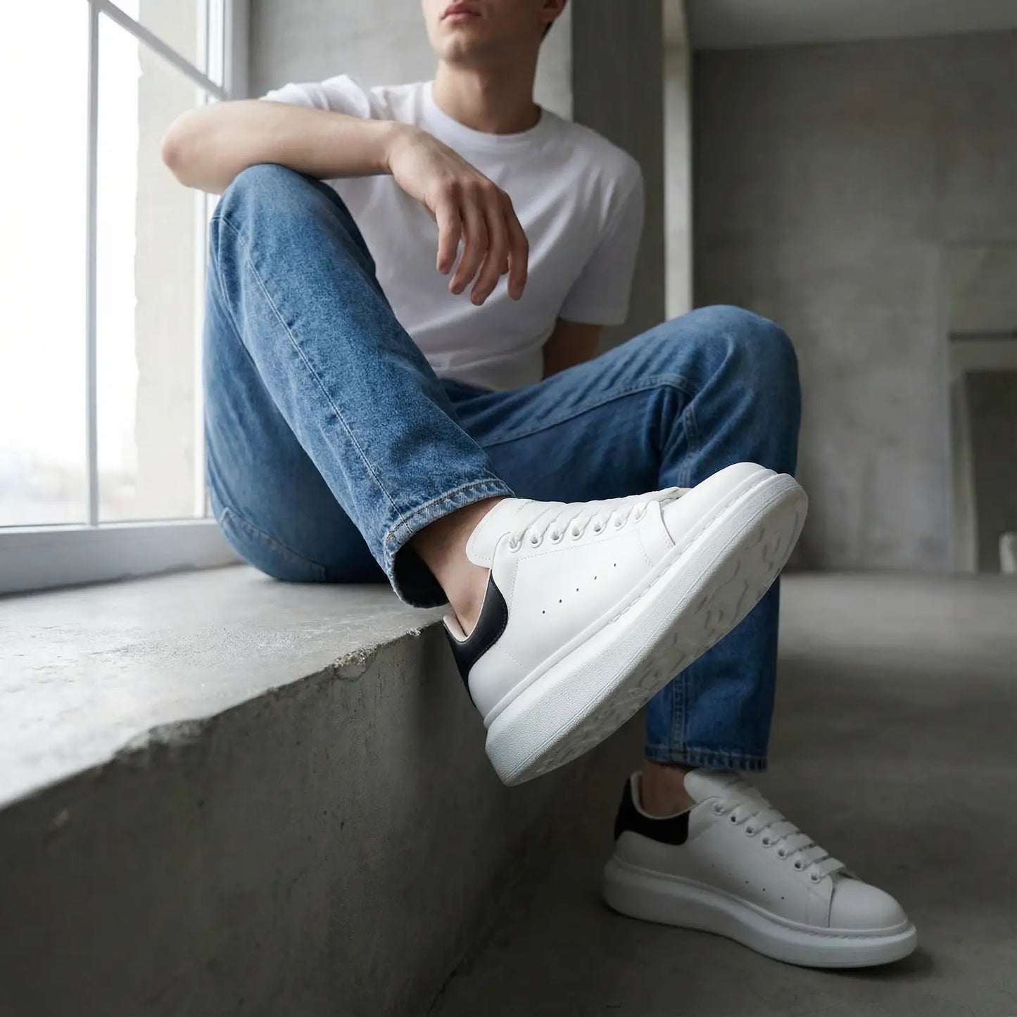 Person wearing white sneakers and blue jeans sitting by a window.